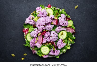 Healthy fresh vegan salad from purple cauliflower, cucumbers, red onion, radicchio, spinach with pumpkin seeds, dark table background, top view - Shutterstock ID 2394909467
