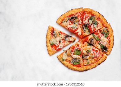 Healthy, gluten free cauliflower crust pizza with tomatoes, mushrooms and spinach. Overhead view with cut slices on a white marble background.