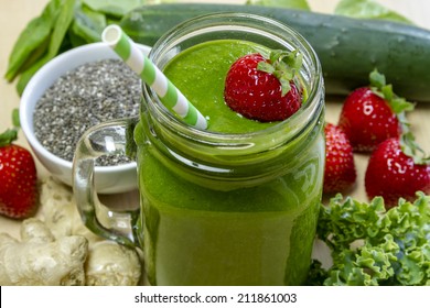 Healthy Green Juice Smoothie Surrounded By Whole Fruits, Vegetables And Chia Seeds With Fresh Strawberry Garnish And Green Swirl Straw