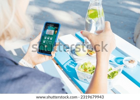 Healthy habit to drink water. Close up woman with glass bottle of pure water with lemon checking phone while sitting at picnic table. Using mobile app to control body hydration, Track water balance