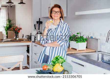 Healthy habit to drink water. Smiling middle age woman with glass of pure water with lemon standing on her kitchen. Control body hydration, Track water balance. Healthy living. Selectivbe focus