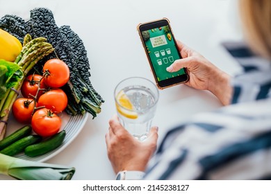 Healthy habit to drink water. Top view Middle age woman with glass of pure water with lemon checking phone while sitting on her kitchen. Using mobile app to control body hydration, Track water balance