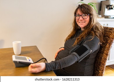 A Healthy And Happy Woman Is Seen In The Third Trimester Of Pregnancy, Sitting At A Dining Table With A Blood Pressure Monitor And Inflatable Arm Cuff