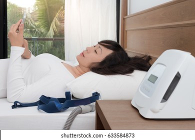  
Healthy And Happy Woman Using Smart Phone On Bed 
With CPAP Machine And Mask In Front.Obstructive Sleep Apnea Therapy.