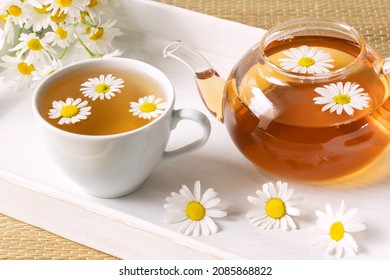 Healthy herbal tea in cup, chamomile flowers and teapot on white wooden tray.