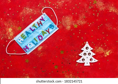 Healthy Holidays Christmas And New Year Concept Banner During Coronavirus COVID-19 Time. Top View Of Disposable Protective Face Mask With Letters On Red Glittered Background