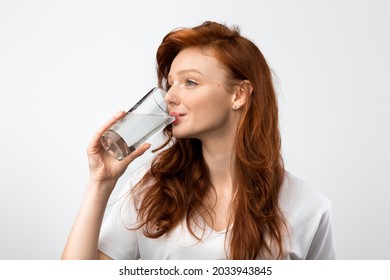Healthy Hydration. Red-Haired Millennial Woman Drinking Water From Glass Standing Over Gray Studio Background, Wearing Casual T-Shirt. Side View Shot. Stay Hydrated Concept