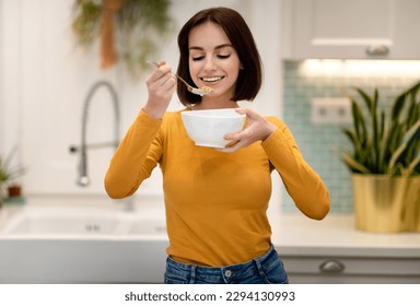 Healthy joyful pretty short-haired young lady in casual outfit holding white bowl and spoon, happy woman having breakfast at home, eating granola, oatmeal or cereals, copy space - Powered by Shutterstock