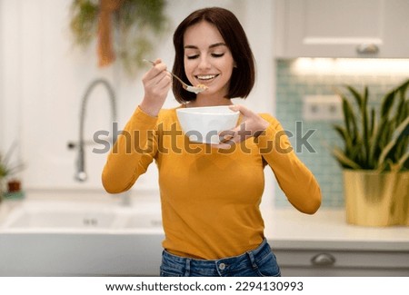 Healthy joyful pretty short-haired young lady in casual outfit holding white bowl and spoon, happy woman having breakfast at home, eating granola, oatmeal or cereals, copy space
