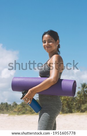 healthy latin brazilian woman dressed in sports clothes with a yoga mat on the beach