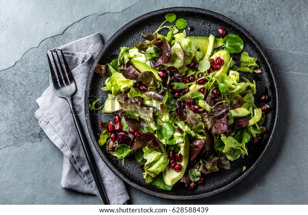 Healthy lettuce salad\
with watercress salad, avocado and pomegranate on black plate. Gray\
slate background