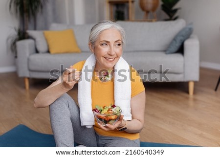 Healthy lifestyle concept. Happy senior woman eating fresh vegetable salad, sitting on yoga mat after home workout, free space. Mature lady keeping weight loss diet