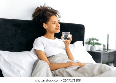Healthy lifestyle concept. Positive, pretty african american young woman, lying in bed in the morning, holds a glass of clean water, smiles, cares of her health, drinks the daily norm of water
