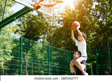 Healthy Lifestyle. Curly son sitting on his daddy's shoulders, throwing ball into basket on sunny day - Powered by Shutterstock