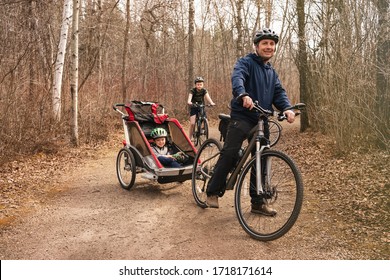 Healthy Lifestyle - Family Biking In Forest Or Park.People Are Riding Bicycles In City Park During Spring Or Early Fall Time. Man Pulls The Sport Trailer Stroller By His Bike With Two Sons. Happiness.