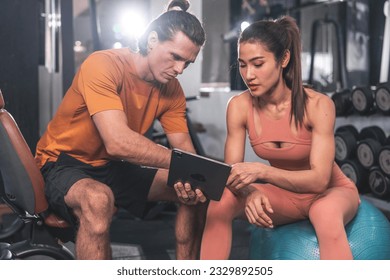 Healthy lifestyle and fitness concept. Asian woman wearing sports bra sitting on yoga ball looking at exercise schedule tablet with Caucasian male trainer in gym. - Powered by Shutterstock