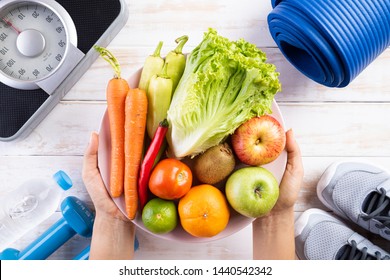 Healthy lifestyle, food and sport concept. Top view of woman hand holding plate of fresh vegetables and fruits with athlete's equipment Weight Scale blue dumbbell, sport water bottles on  wooden. - Powered by Shutterstock