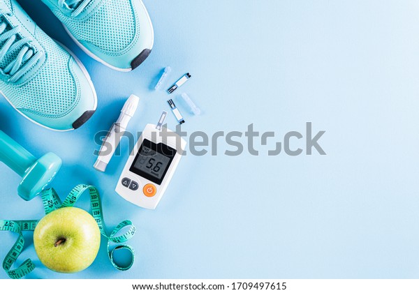 Healthy lifestyle, food\
and sport concept. Top view of diabetes tester set with athlete\'s\
equipment; measuring tape, green dumbbell and  fruit on bright blue\
pastel background.