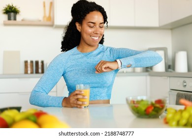 Healthy lifestyle. Happy fit african american woman in sports bra holding fresh juice and looking at wrist smartwatch, sitting at table in kitchen - Powered by Shutterstock