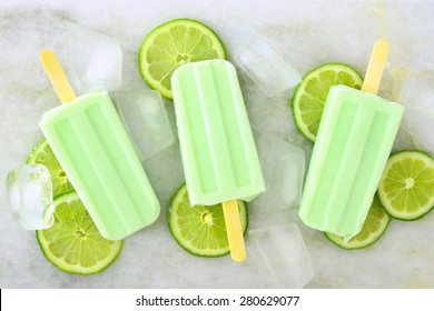 Healthy lime yogurt popsicles with fresh lime slices scattered on a white marble background