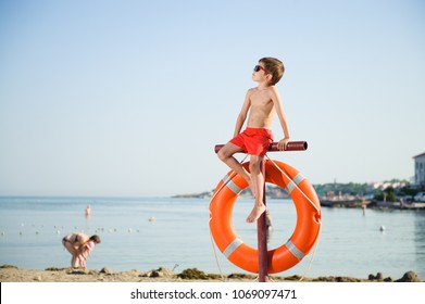 Healthy Little Boy In Sunglasses Sits On Pole With Orange Lifebuoy Hanging On It On Summer Beach With People In Baywatch