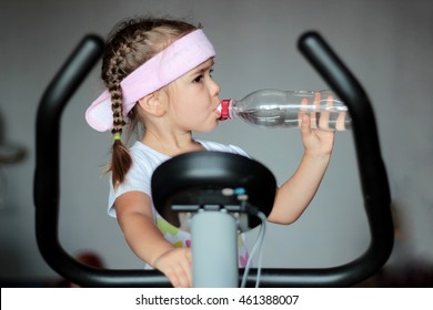Healthy little girl drinking water from the bottle during training at home on exercise bike, healthy lifestyle concept