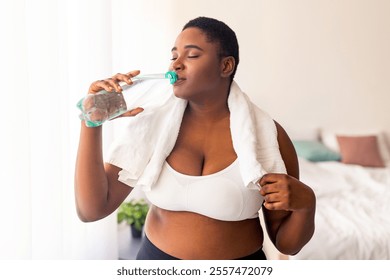 Healthy living, weight loss. Positive plus size black lady drinking fresh clear water from bottle after sports workout indoors. Curvy millennial woman staying hydrated during home fitness exercises - Powered by Shutterstock