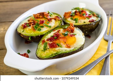 Healthy low carb paleo breakfast of baked avocado and eggs boats with bacon crumbles and chives sitting in white baking dish with two forks