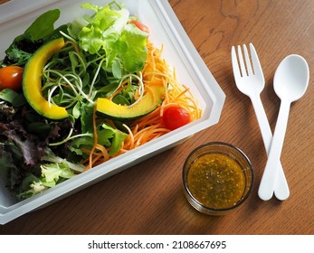 Healthy lunch box preparation : Mixed fresh vegetable with steamed pumpkin salad contained in bioplastic take out packaging on wooden table with honey olive oil dressing, white plastic fork and spoon.