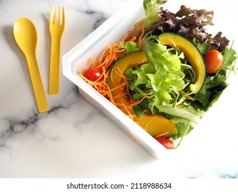 Healthy lunch box preparation : Mixed fresh vegetable with steamed pumpkin salad contained in bioplastic take out packaging on marble table with honey olive oil dressing, yellow plastic fork and spoon