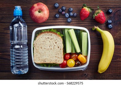 Healthy lunch box with sandwich and fresh vegetables, bottle of water and fruits on wooden background. From top view