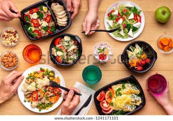Healthy lunch time at office workplace. Four people\
eating healthy meals from take away lunch boxes and plates at\
wooden table. concept, top\
view