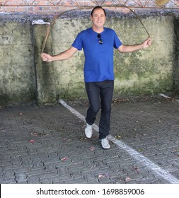 Healthy Man About 50 Years Old Skipping Rope In A Car Garage, Exercising Happily Wearing Jeans, Blue Shirt And Tennis Shoes