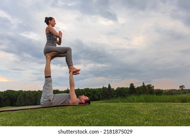 Healthy Man Lying On Grass And Balancing Woman. Couple Doing Acrobatic Yoga Exercise In Park