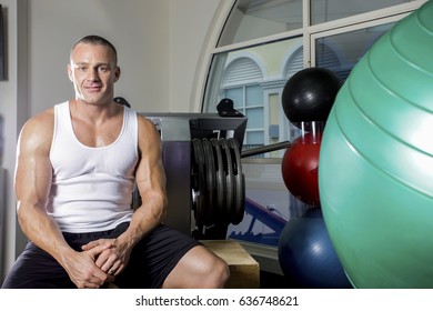 Healthy Man Relaxing After Work Out,he Has A Strong Body, Inside Room Of A Luxury Gym, Heavy Exercise Tools Near Wall, A Body Builder Sitting On A Bench.