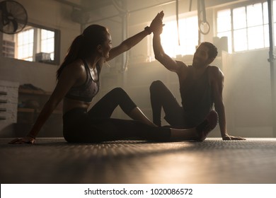 Healthy man and woman sitting on floor and giving each other high five at the gym. Fitness people after successful exercising session in gym.