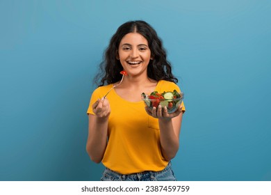 Healthy Meal. Portrait Of Happy Casual Indian Woman Eating Tasty Fresh Vegetable Salad, Holding Plate Bowl And Fork Looking At Camera. Satisfied Young Lady Isolated On Blue Background - Powered by Shutterstock