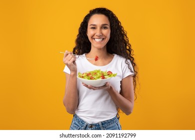 Comida saludable. Retrato De Una Feliz Mujer Casual Latino Comiendo Una Ensalada Fresca De Vegetales, Sujetando Un Tazón De Plate Y Una Horquilla Mirando La Cámara. Dama Del Milenio Satisfecha Aislada En Fondo Naranja Amarillo