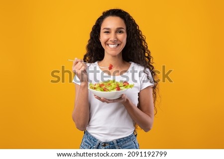 Healthy Meal. Portrait Of Happy Latin Casual Woman Eating Tasty Fresh Vegetable Salad, Holding Plate Bowl And Fork Looking At Camera. Satisfied Millennial Lady Isolated On Yellow Orange Background