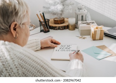 Healthy New Years Resolutions For Older Adults. Senior Mature Old Woman In White Sweater Writing Handwritten Text New Years Resolutions In Open Notepad On The Table. Planning And Setting Goals