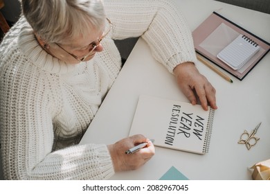 Healthy New Years Resolutions For Older Adults. Senior Mature Old Woman In White Sweater Writing Handwritten Text New Years Resolutions In Open Notepad On The Table. Planning And Setting Goals