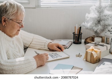 Healthy New Years Resolutions For Older Adults. Senior Mature Old Woman In White Sweater Writing Handwritten Text 2022 Goals In Open Notepad On The Table. Planning And Setting Goals