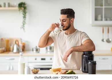 Healthy nutrition for muscle gain and weight loss concept. Athletic young Arab man drinking protein shake or milk, standing near table with healthy wholesome products at kitchen, copy space