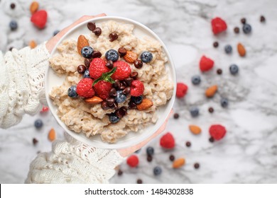 Healthy oatmeal served with berries, chocolate chips, almonds and honey. Bowl held in a womans hands over a marble table background. Shot from top view.
