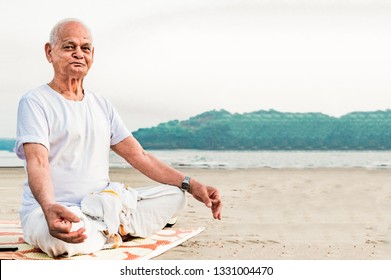 Healthy Old Indian Man, Super Senior Citizen Performing Yoga, Sitting In Lotus Pose And Doing Meditation On Beach. He Is Wearing Dhoti, Has Pretty Wrinkles On His Face And Is Happily Posing For Camera