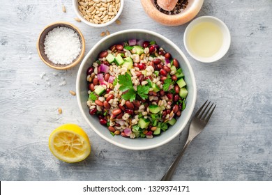 Healthy pearl barley salad with beans, cucumbers, red onion, sunflower seeds, pomegranate and parsley in bowl on concrete background. Top view.
