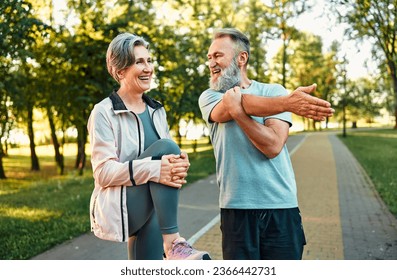 Healthy pensioners exercising outside. Smiling man and woman of senior age standing on paved road and doing physical activities. Slim lady lifting and clutching knee to belly while man stretching arm. - Powered by Shutterstock