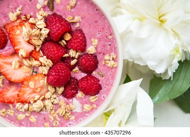 Healthy Pink Smoothie in the Bowl from Banana and Strawberries with Pieces of Strawberries, Raspberries, Granola and Coconut Shreds on top. Peonies nearby. Top View - Powered by Shutterstock