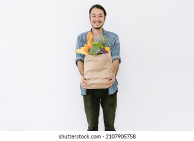 Healthy Positive Young Asian Man Holding Paper Shopping Bag Full Of Fresh Vegetables And Groceries With Copy Space Isolated On White Background. Happy Japanese Or Korean Male Buyer Came From Market.