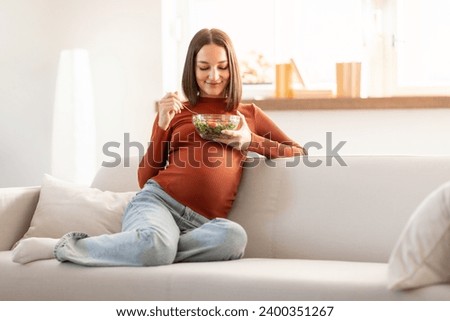 Healthy Pregnancy Nutrition. Glad Pregnant Woman Eating Fresh Veggie Salad While Sitting On Sofa Indoor, At Modern Home Interior. Holding Bowl With Tasty Organic Food, Enjoying Expectation Menu